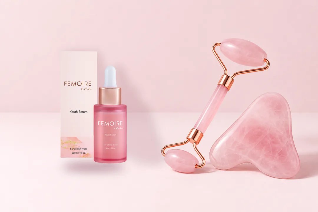 淺粉紅色枱面上嘅一套 FEMOIRE：青春精華液同盒、粉晶滾輪同心形刮痧板