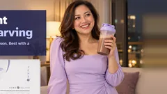 A confident woman in a lavender dress smiling with her chocolate Kuro Daitto shake, the Kuro Daitto box on the side table. Lose weight without starving. Results vary from person to person. Best with a balanced diet and exercise.