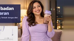 Seorang wanita yakin berpakaian ungu lembut tersenyum dengan bancuhan Kuro Daitto coklat, kotak Kuro Daitto di atas meja sisi. Turun berat badan tanpa kelaparan. Hasil berbeza bagi setiap orang. Terbaik dengan diet seimbang dan senaman.