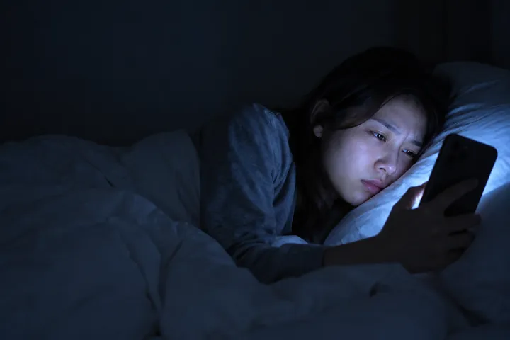 Illustration: a woman lying in bed at night, her face lit by the cold blue glow of her phone