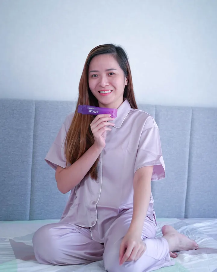 A woman in pyjamas on her bed, smiling, a RELAZZ sachet in her hand